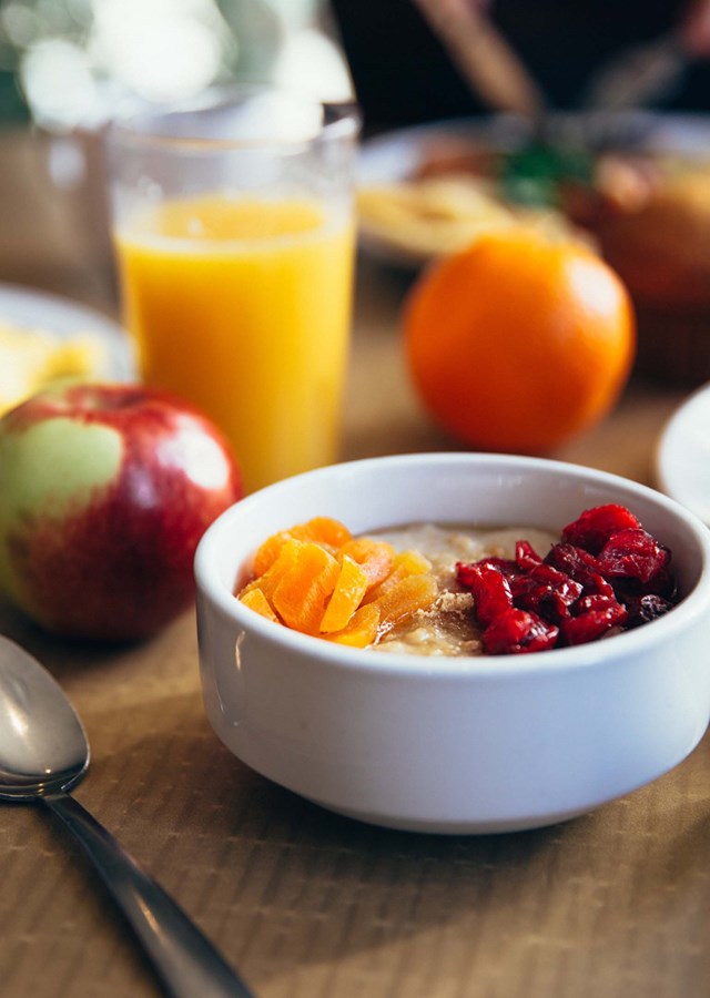 A white bowl full of fruit with a spoon next to it sitting on a table. The rest of the table contains more fruit and other healthy breakfast foods  