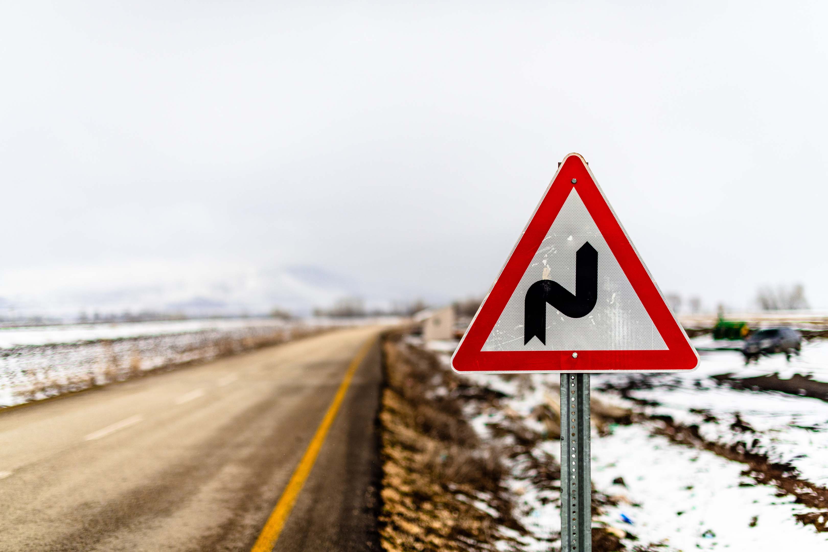 A road warning sign showing bends ahead next to a road with snow covered fields on either side
