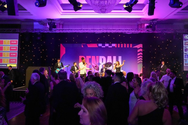 Guests in black tie fill the dance floor as a live band performs on stage in front of a Ben Ball backdrop, with auction bids displayed on a screen.