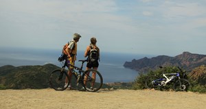 A couple of cyclists with bicycles standing on cliff top over looking a beach and rocky outcrop reaching out in to the sea