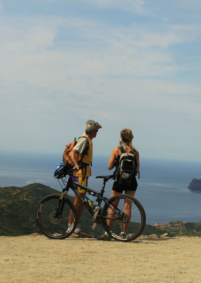 A couple of cyclists with bicycles standing on cliff top over looking a beach and rocky outcrop reaching out in to the sea