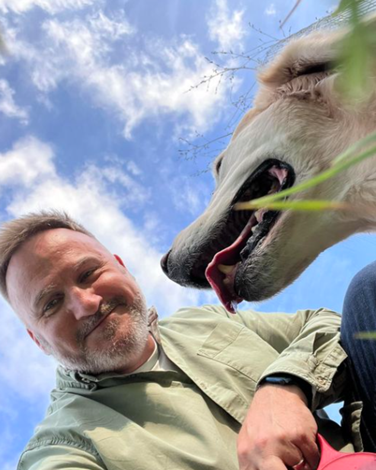 A smiling man with a grey beard outdoors with his large white dog, taken from below against a blue sky.
