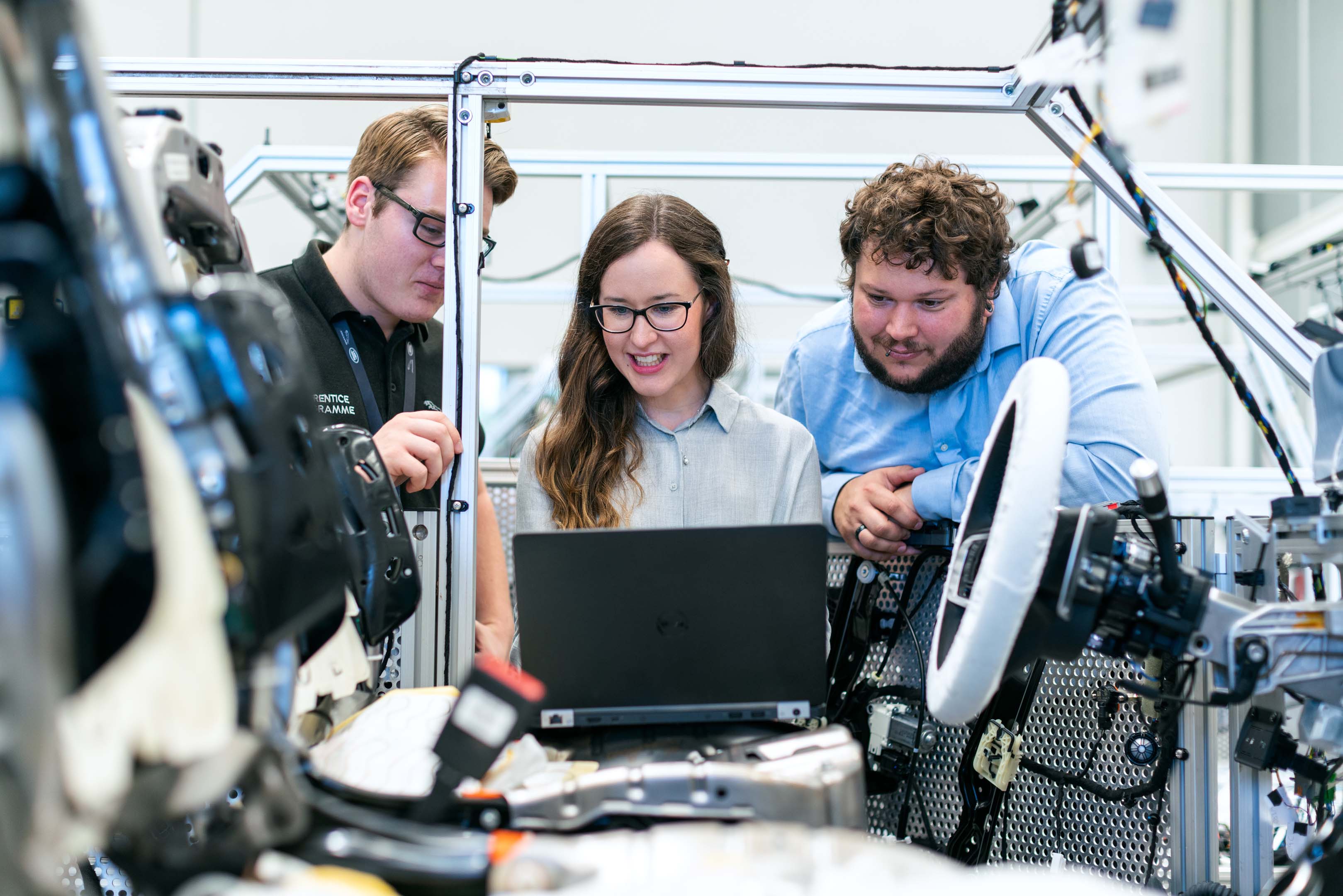 Three colleagues in an engineering environment standing & working together with a laptop connected to a vehicle 