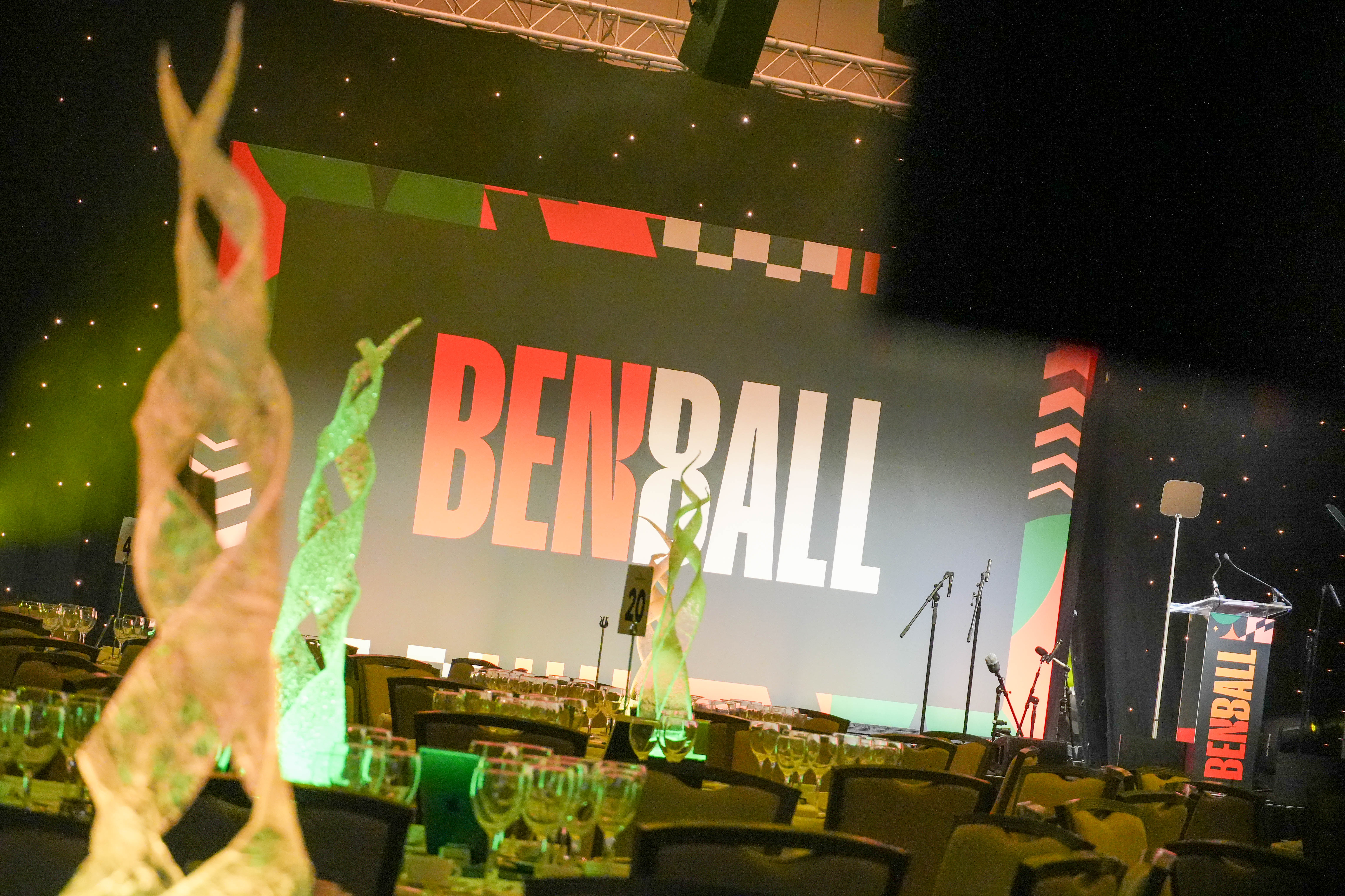 Ben Ball venue set up before guests arrive, with a large 'Ben Ball' stage screen, lit table centrepieces and rows of set tables.