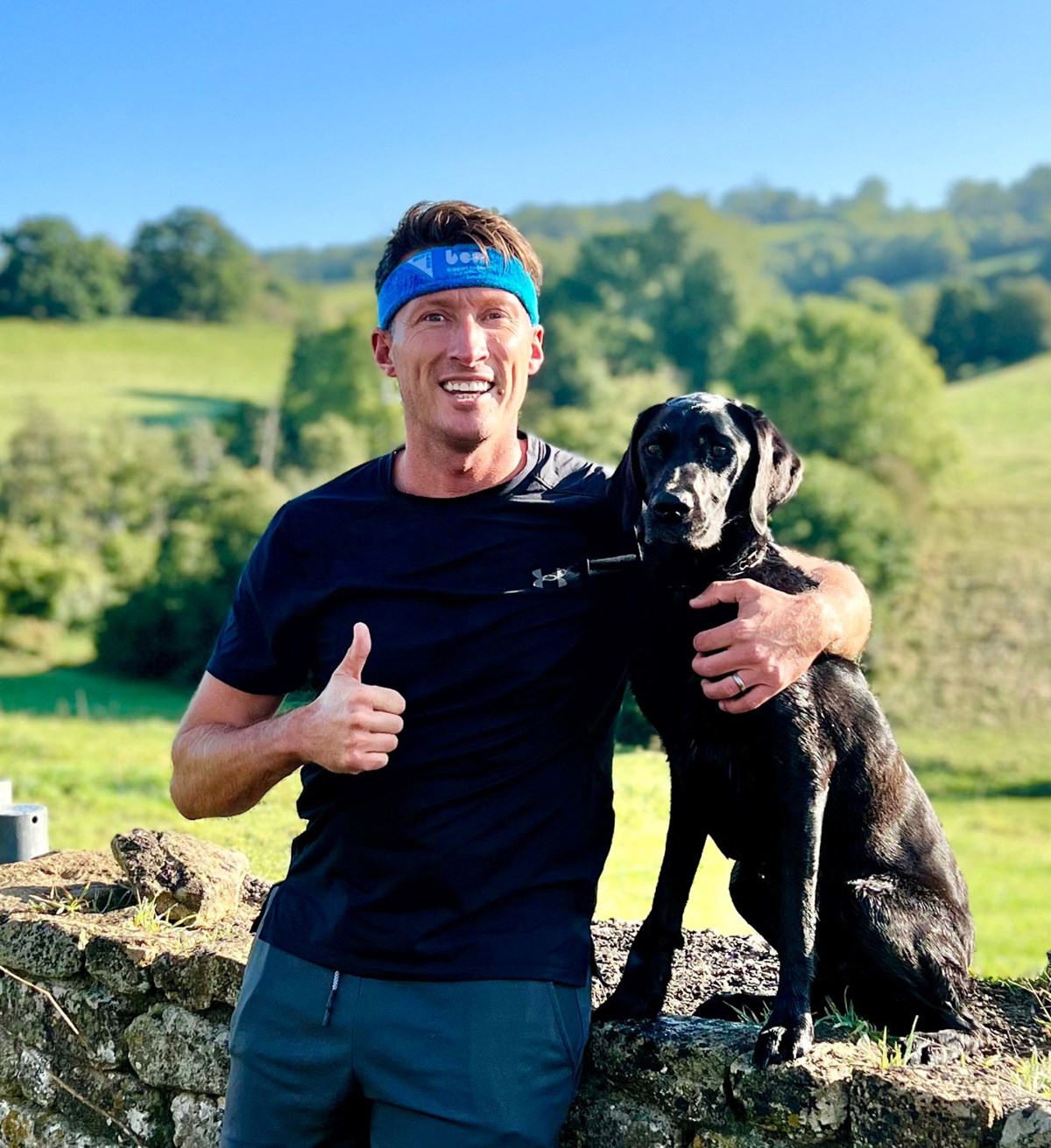 Man in a blue Ben headband and black sportswear gives a thumbs up beside a black dog sitting on a stone wall in the countryside.