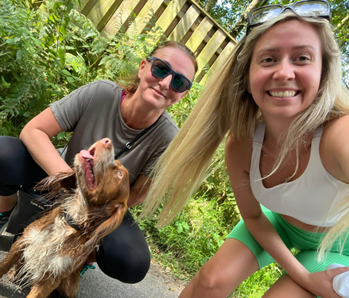 Two women crouch down for a selfie outdoors on a sunny path with a happy spaniel between them.