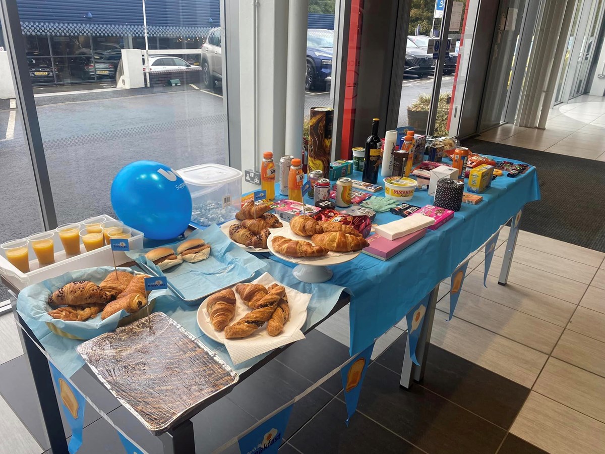 A Big Breakfast fundraising table in a car dealership showroom, laid with croissants, pastries, rolls, orange juice, drinks and raffle prizes, decorated with blue bunting and a balloon.