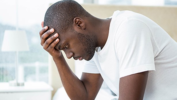 A man sits on the edge of a bed with his head bowed and his hand on his forehead, looking distressed.