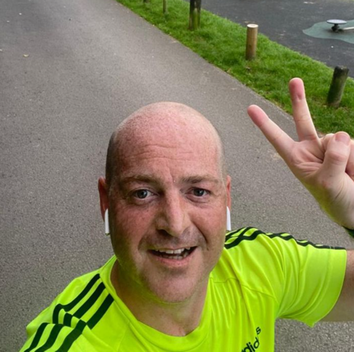 Man in a hi-vis yellow running top taking a selfie on a park path, making a peace sign.