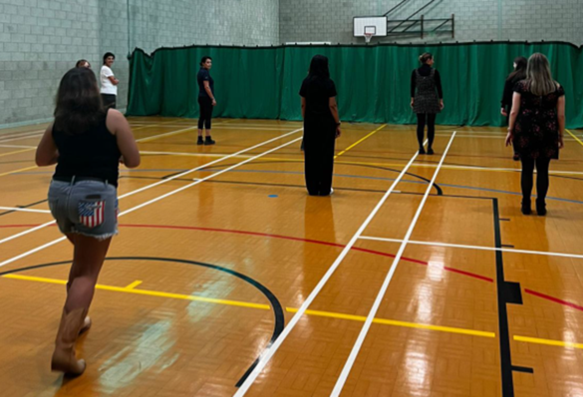 A group of women standing spaced apart in a sports hall, taking part in an indoor exercise session.