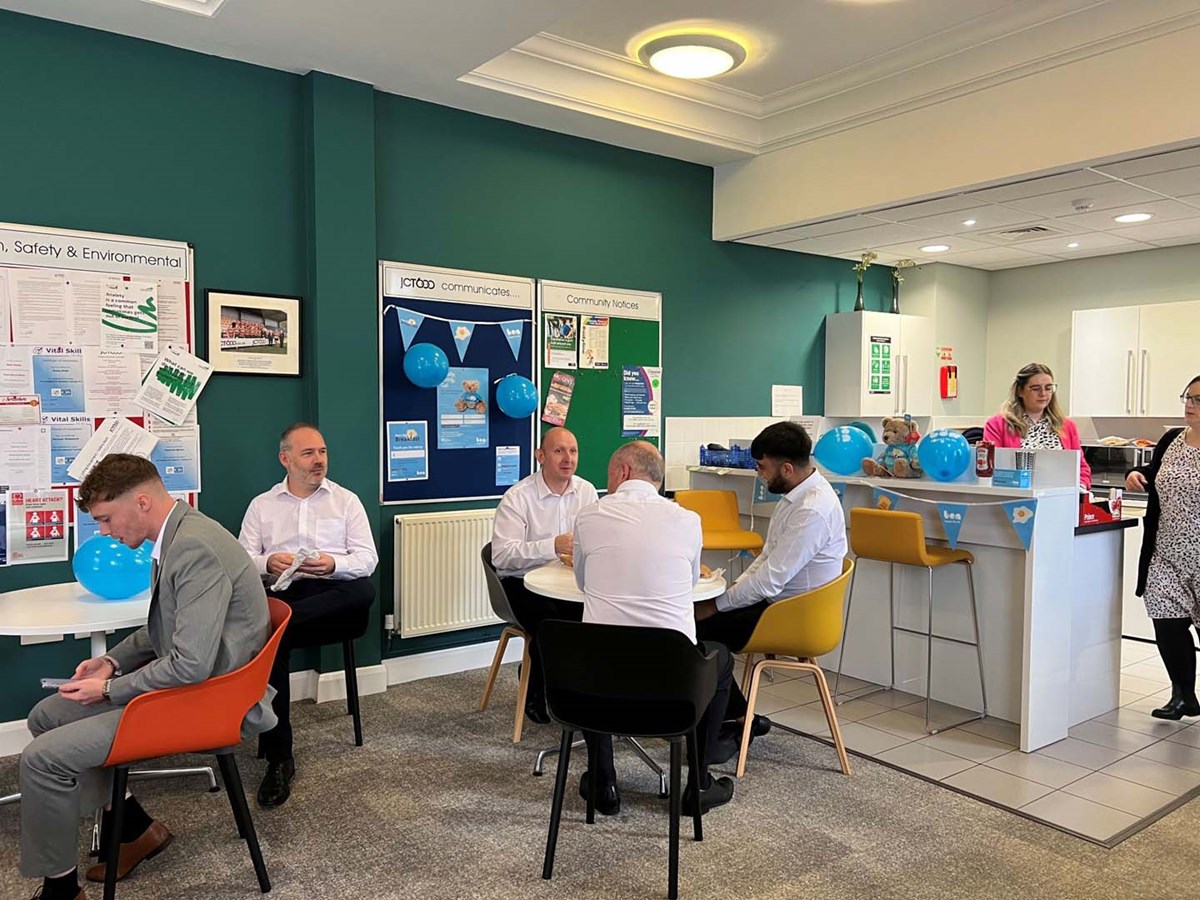 Colleagues sitting at tables in a workplace kitchen decorated with blue Ben balloons and bunting for Ben's Big Breakfast.